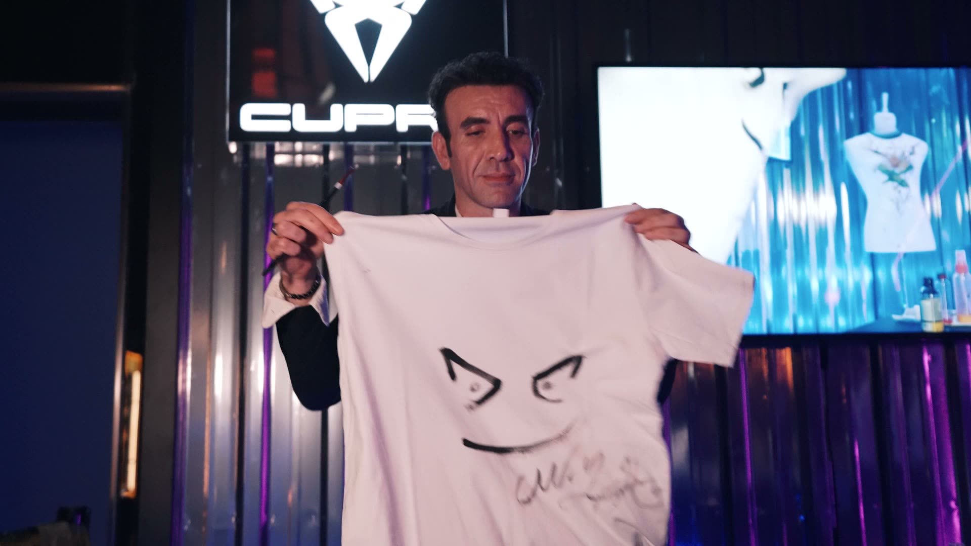 A man holding up a printed shirt in front of a Cupra backdrop